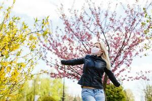 gelukkig jong vrouw vervelend gelaats masker voor virus bescherming staand buitenshuis Aan zonnig voorjaar dag. foto