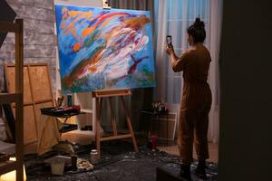 schilder nemen foto gebruik makend van smartphone van groot schilderij in kunst studio. modern artwork verf Aan canvas, creatief, hedendaags en geslaagd prima kunst artiest tekening meesterwerk