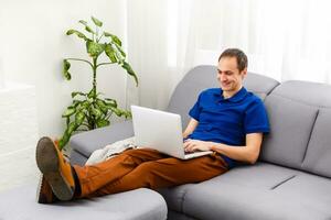gelukkig jong Mens in t-shirt zittend Bij huis, werken Aan laptop computer, lachend. foto