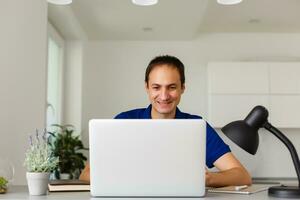 gelukkig jong Mens in t-shirt zittend Bij huis, werken Aan laptop computer, lachend. foto