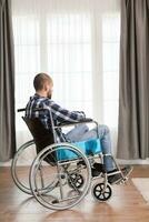 bezorgd jong Mens in rolstoel leven alleen echt verdrietig Kaukasisch Mens vervelend gewoontjes kleren en kijken Bij groot panoramisch visie in helder modern leven kamer, ziekenhuis of kliniek. foto