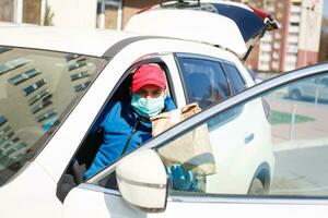 levering Mens Holding papier zak met voedsel in de buurt de auto, voedsel levering Mens in beschermend masker foto