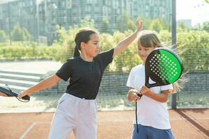 tiener- meisjes met rackets en ballen staand in padel rechtbank, op zoek Bij camera en lachend. foto