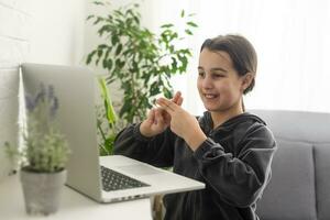 glimlachen tiener- meisje doof gehandicapt kind school- meisje aan het leren online klasse Aan laptop communiceren met leraar door video conferentie telefoontje gebruik makend van teken taal tonen hand- gebaar gedurende virtueel les. foto