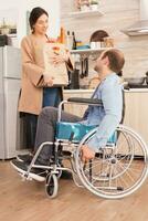 vrouw Holding papier zak met biologisch producten in keuken pratend met gehandicapt man in rolstoel. gehandicapt verlamd gehandicapten Mens met wandelen onbekwaamheid integreren na een ongeluk. foto