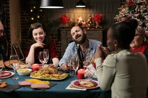 blij mensen genieten van winter feest Bij tafel, jong en oud volwassenen hebben pret aan het eten eigengemaakt maaltijden en drinken wijn. vrienden en familie bijeenkomst in de omgeving van voor feestelijk Kerstmis avondeten feest. foto