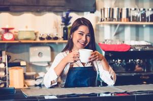 Aziatische vrouwelijke barista kopje koffie maken. jonge vrouw die een witte koffiekop vasthoudt terwijl ze achter de cafébar op de achtergrond van het restaurant staat. mensenlevensstijlen en bedrijfsbezettingsconcept