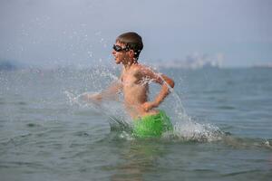 een gelukkig jongen loopt aan de overkant de zee spatten water. de kind baadt, zonnebaadt en rust. zomer vakantie. foto