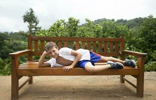 een gelukkig kind in de zomer warmte leugens Aan een houten bank tegen de backdrop van tropisch planten. de jongen is Aan vakantie in de zomer. foto