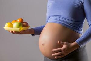 beeld van dichtbij omhoog maag van zwanger vrouw Holding fruit Aan grijs achtergrond. foto