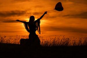 silhouet van een vrouw zittend Aan koffer met hart vormig ballon en op zoek Bij mooi zonsondergang. foto