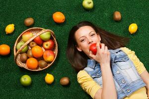 mooi jong vrouw is aan het eten appel terwijl aan het liegen naar beneden Aan de gras met een veel van fruit in de omgeving van haar foto