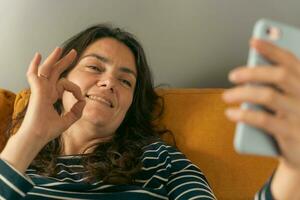 positief middelbare leeftijd vrouw maken video telefoontje tonen OK gebaar Aan smartphone foto