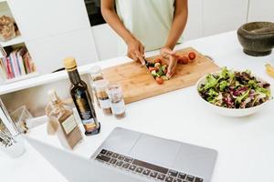 zwarte jonge vrouw die salade maakt terwijl ze laptop in de keuken gebruikt