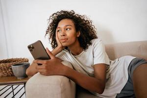 zwarte jonge vrouw die mobiele telefoon gebruikt terwijl ze op de bank rust foto