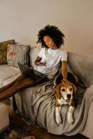 zwarte jonge vrouw die mobiele telefoon gebruikt en haar hond op de bank aait