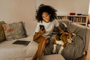 zwarte jonge vrouw die mobiele telefoon gebruikt en haar hond op de bank aait
