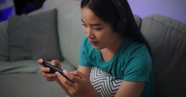 portret van jong Aziatisch vrouw vervelend bril en hoofdtelefoons geniet spelen online esport spellen Aan smartphone zittend Aan sofa in de leven kamer Bij thuis, gamer levensstijl concept. foto