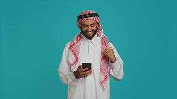 moslim persoon in traditioneel kostuum hebben smartphone in handen en typen in internet sociaal media berichten. jong volwassen browsen Aan mobiel telefoon en sms'en, internet webpagina. foto