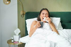 Gezondheid en mensen. Aziatisch meisje aan het liegen in bed, gevoel ziek, niezen en drinken heet thee, vangen koud, blijven Bij huis Aan vertrekken foto