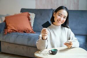 jong vrouw in leven kamer, tonen haar credit kaart en gebruik makend van smartphone naar betalen, bestellen online of boodschappen doen Aan toepassing foto