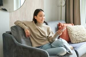 portret van modern vrouw zittend Bij huis met op afstand, ontspannende Aan weekenden, aan het kijken TV televisie en glimlachen foto