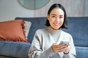 mensen en technologie. glimlachen Aziatisch vrouw zittend Bij huis, gebruik makend van haar mobiel telefoon, typen bericht, wenkbrauwen internet of boodschappen doen online van smartphone app foto