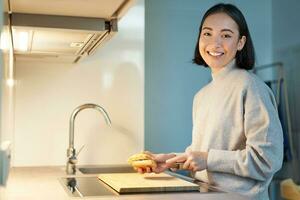 portret van glimlachen jong Aziatisch vrouw staand Aan keuken en maken een sandwitch, Koken voor haarzelf foto