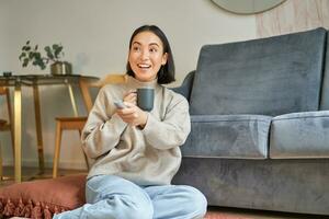 portret van glimlachen jong vrouw resting Bij huis in de buurt TV, aan het kijken televisie zittend Aan verdieping en drinken koffie van kop foto