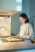verticaal schot van jong Aziatisch vrouw Koken diner, maken haarzelf sandwitch, glimlachen terwijl staand Aan de keuken foto