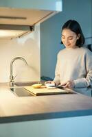 verticaal schot van jong Aziatisch vrouw Koken diner, maken haarzelf sandwitch, glimlachen terwijl staand Aan de keuken foto