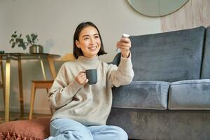 portret van glimlachen Koreaans vrouw zittend in de buurt TV, Holding afgelegen en schakelen kanalen terwijl drinken heet koffie foto