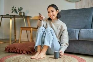 portret van mooi Aziatisch meisje zittend Bij haar huis en aan het kijken TV, Holding op afstand, glimlachen en lachend, gevoel comfort en warmte Bij haar appartement foto
