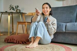 beeld van jong Koreaans vrouw drankjes koffie, houdt afgelegen van TV, aan het kijken televisie Bij huis, resting in haar huis foto