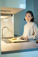 verticaal schot van jong Aziatisch vrouw Koken diner, maken haarzelf sandwitch, glimlachen terwijl staand Aan de keuken foto