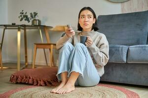 portret van jong vrouw met op afstand, aan het kijken TV, schakelen kanalen Aan televisie, zittend Aan verdieping in de buurt sofa en ontspannende foto