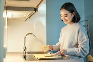 portret van glimlachen jong Aziatisch vrouw staand Aan keuken en maken een sandwitch, Koken voor haarzelf foto