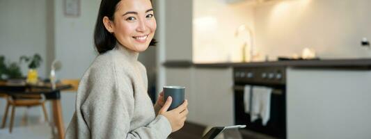 portret van glimlachen Aziatisch meisje met kop van koffie, drinken heet thee en krijgen knus Bij huis. opwarming omhoog met drank foto