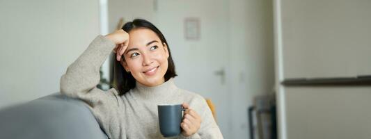 glimlachen Aziatisch vrouw zittend Aan sofa met haar mok, drinken koffie Bij huis en ontspannende na werk, op zoek kalmte en knus foto