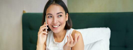 dichtbij omhoog portret van schattig Aziatisch meisje in bed, pratend Aan mobiel telefoon met gelukkig glimlachen gezicht. vrouw ontwaken omhoog en maken een telefoon telefoontje foto