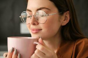 dichtbij omhoog portret van mooi glimlachen vrouw, drankjes haar ochtend- kop van koffie, geniet smakelijk aroma, ruiken haar drinken met verheugd gezicht foto