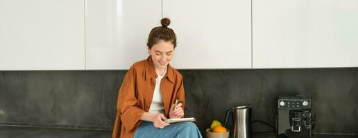 portret van elegant jong vrouw, zit Bij huis in de keuken en maakt notities, schrijft naar beneden recept, cheques haar lijst met huis taken, doodling of tekening in notitieboekje foto