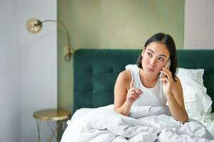 mooi jong Koreaans vrouw roeping, hebben een telefoon telefoontje in bed, pratend met vriend terwijl blijven in slaapkamer foto