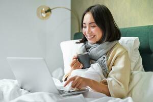 portret van jong Aziatisch vrouw aan het liegen ziek in bed, blijven Bij huis met verkoudheid of griep, aan het kijken videos, gebruik makend van laptop in haar slaapkamer foto