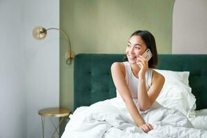 mooi glimlachen Aziatisch meisje pratend Aan mobiel telefoon, aan het liegen in bed met verheugd gelukkig gezicht, sprekend naar iemand Aan telefoon foto