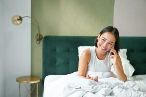 glimlachen Koreaans meisje praat Aan mobiel telefoon en aan het liegen in bed. schattig vrouw antwoorden telefoon telefoongesprek, houdt smartphone, ontspannende in haar bed foto