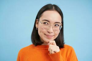 portret van attent Koreaans vrouw in bril, denken en op zoek terzijde, heeft een idee, plannen iets, staat over- blauw achtergrond foto