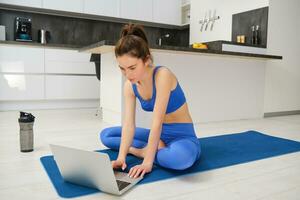 beeld van jong geschiktheid instructeur gebruik makend van laptop voor online training opleiding, tonen geschiktheid opdrachten, pratend naar cliënt, zittend Aan yoga rubber mat in sportkleding foto