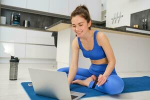 portret van jong vrouw in aansluiting op online geschiktheid les Bij huis, zittend Aan yoga mat en gebruik makend van laptop gedurende aerobics training foto