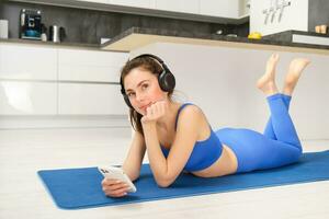 portret van mooi geschiktheid meisje, houdende Aan yoga mat, gebruik makend van mobiel telefoon en draadloze koptelefoon, op zoek attent en glimlachen foto